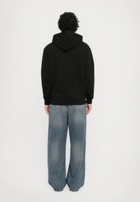 Hanorac negru cu glugă ajustabilă, purtat împreună cu blugi oversized, de un albastru deschis. Țesătura pare moale, cu o croială lejeră și un design simplu.