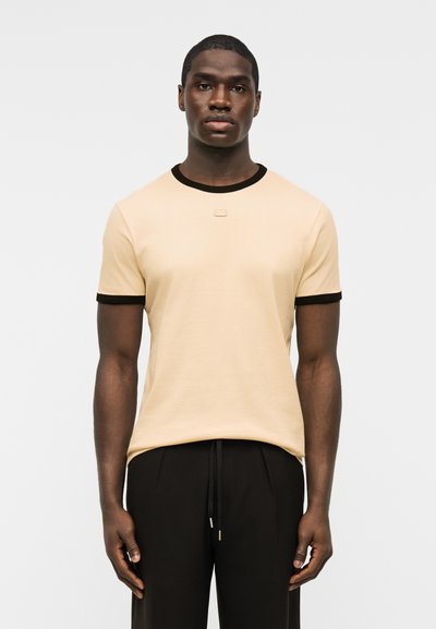 Άνδρας όρθιος, φοράει μπεζ κοντομάνικο μπλουζάκι με μαύρο γιακά και άκρα μανικιών, σε συνδυασμό με μαύρο παντελόνι, ουδέτερο φόντο.