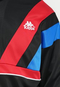 Černá bunda s červenými a modrými diagonálními pruhy. Obsahuje bílé lemování podél spodního lemu a bílé logo Kappa na hrudi. Texturovaná tkanina.