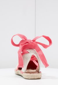 Sandalia de espadrille con suela de yute trenzado, parte superior de tela en tono marrón claro y rosa, con un diseño de apertura trasera y largas cintas de tela rosa para atar.