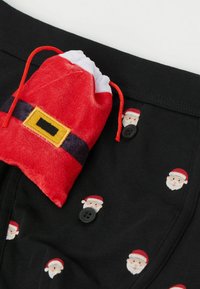 Czarne szorty z tkaniny z powtarzającym się nadrukiem twarzy Świętego Mikołaja. Zawiera mały czerwony welurowy woreczek z żółtym paskiem.