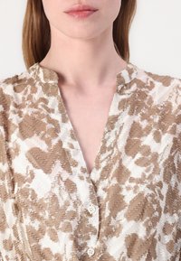 Blusa stampata beige e bianca con una placca di bottoni visibile, texture morbida e scollo a V. Il tessuto presenta un motivo sottile e testurizzato.