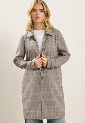 Kvinde iført en ternet frakke i mellemlang længde med brune knapper over en hvid skjorte, der holder frakken lukket med begge hænder.