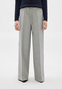 Persona, kas valkā gaiši pelēkas plašas bikses ar augstu vidukli un aukliņu, kā arī tumši zilu džemperi, stāv pret vienkāršu fonu.