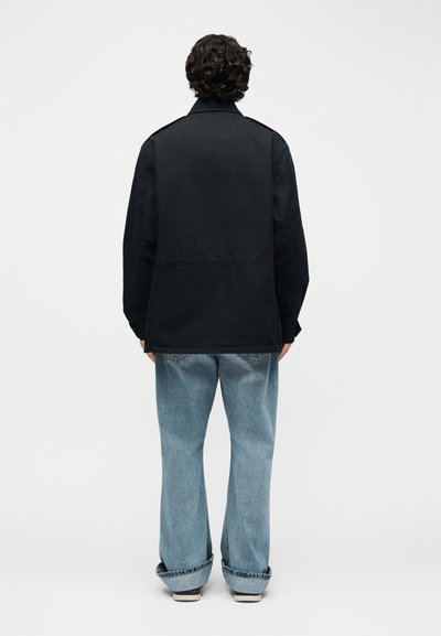 Άτομο με σγουρά μαλλιά που φοράει μαύρο σακάκι και ανοιχτό μπλε φαρδιά τζιν, στέκεται με το πρόσωπο στραμμένο προς ένα απλό λευκό φόντο.