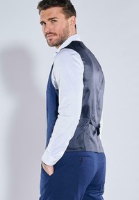 Marineblaue Weste mit satinierter Rückseite, tailliertem Schnitt, zwei Fronttaschen und Knopfverschluss. Wird über einem weißen Hemd getragen.