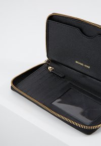 Portefeuille noir Michael Kors avec fermeture éclair tout autour, ouvert pour montrer les emplacements pour cartes, une fenêtre d'identification transparente et des détails dorés sur la fermeture éclair.