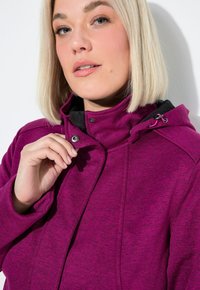 Kobieta z blond bobem, nosząca fioletową kurtkę z kapturem, zapięta na nap przy kołnierzu na jednolitym białym tle.