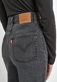 Gros plan sur une personne portant un jean noir Levi's avec un patch en cuir de la marque visible et une étiquette rouge Levi's sur la poche arrière.