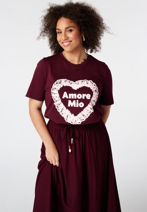 Glimlachende vrouw met krullend haar, draagt een bordeauxkleurig shirt en rok; het shirt heeft een wit hart met de tekst "Amore Mio" erin.