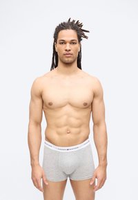 Μυώδης άνδρας με κοντούς κοτσίδες στέκεται κοιτώντας προς τα εμπρός φορώντας γκρι boxer briefs Tommy Hilfiger σε απλό ανοιχτόχρωμο φόντο.