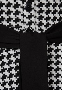 Svart och vit mönstrad kofta med houndstooth-design, knappstängningar och ett knytbart svart tygbälte i midjan.