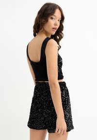 Joven posando con shorts negros de lentejuelas y un top corto sin mangas a juego, sobre un fondo blanco liso.