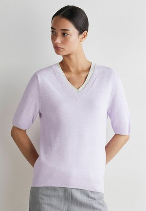 Ung kvinde iført en lys lilla kortærmet V-hals sweater og grå bukser, stående med hænderne bag ryggen mod en hvid væg.