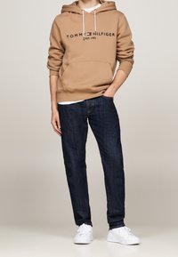 Felpa marrone con logo "TOMMY HILFIGER", tasche anteriori e coulisse bianche, abbinata a jeans blu scuro e sneakers bianche.