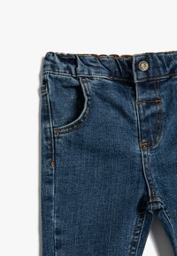 Vaqueros de denim en un color azul oscuro, con cinturilla elástica, bolsillo frontal y costuras visibles en un tono naranja contrastante.