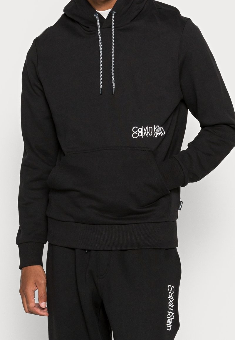Sweat à capuche noir avec une poche avant, doté d'un cordon gris et d'un logo blanc sur le côté droit. Tissu doux et texturé.