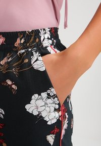 Shorts noirs à motif floral avec une taille élastique et une poche latérale, ornés de fleurs blanches et rouges ainsi que de papillons sur un tissu texturé.