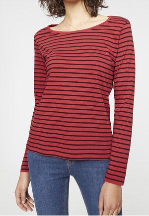 Femme portant un chemisier rouge à manches longues avec des rayures noires horizontales et un jean bleu, debout contre un fond blanc uni.