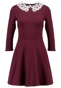 Burgundy pitkähihaineen mekko, jossa on levenevä hame ja valkoinen pitsinen Peter Pan -kaulus.