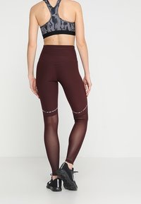Leggings alto color granata con pannelli in rete nella parte bassa delle gambe, abbinati a un reggiseno sportivo a motivi neri e grigi e scarpe da ginnastica nere.