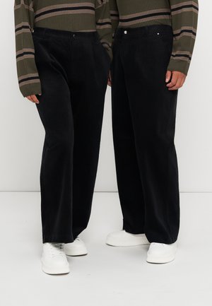 Deux personnes portant un pantalon noir à jambes larges, des hauts à manches longues rayés vert olive et des baskets blanches, debout côte à côte.