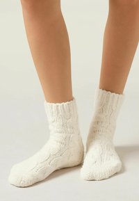 Chaussettes blanches en tricot avec un motif de cable, dotées d'un bord côtelé et d'une texture douce, conçues pour s'ajuster parfaitement autour des chevilles.