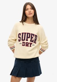 Suéter crema con el logo rojo y azul marino "SUPER DRY" en la parte frontal; puños acanalados con rayas rojas y azules; combinado con una falda plisada azul marino.