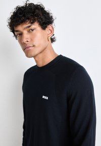 Svart stickad tröja med rund halsringning, med en texturerad design och en liten vit "BOSS"-logotyp på bröstet.
