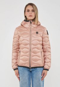 Donna che indossa un giubbotto imbottito rosa chiaro con cappuccio e jeans blu, in piedi contro uno sfondo bianco semplice, rivolta in avanti.