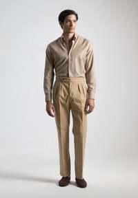 Camisa beige de cuello, de mangas largas, con botones en el frente; pantalones beige con pliegues y una cintura alta, con detalle de un solo botón; zapatos marrones.