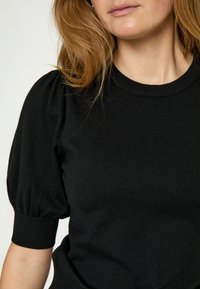Vrouw draagt een zwart gebreide top met korte mouwen, opgeblazen mouwen en geribbelde manchetten, getoond van nek tot taille tegen een effen achtergrond.