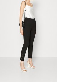 Schwarze, eng anliegende Hosen mit seitlichen Schlitzen und metallischen Akzenten am Saum, kombiniert mit einem weißen Satin-Camisole und leichten Keilsandalen.