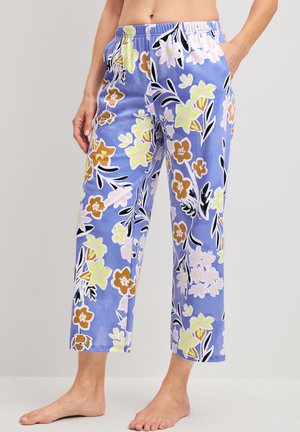 Persoon die blauwe brede bloemenbroek met elastische tailleband draagt, staat blootsvoets tegen een effen achtergrond.