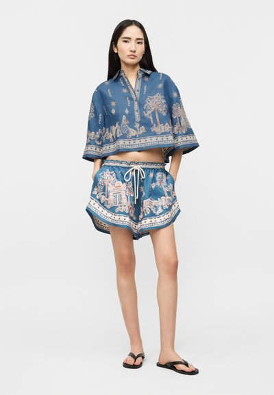 Γυναίκα που φοράει μπλε τοπ και σορτς με μπεζ περίτεχνα σχέδια και μαύρα σαγιονάρες, στέκεται με τα χέρια στις τσέπες σε απλό φόντο.