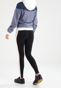 Lättviktsjacka med huva i grått med marinblå och vita detaljer, kombinerad med svarta leggings med en rosa sidremsa. Svarta sneakers.