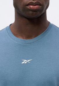 Gros plan d'une personne portant un t-shirt bleu clair avec un petit logo blanc centré sous l'encolure.