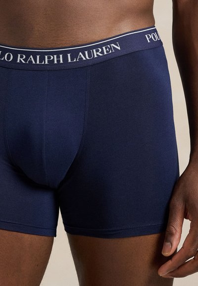 Vyras vilki tamsiai mėlynas „Polo Ralph Lauren“ bokserines trumpikes su logotipo juosmens juosta, stovintis su ranka atsipalaidavusia šalia kūno.