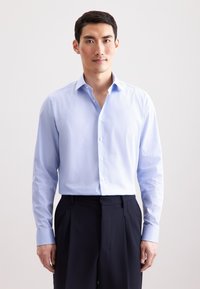 Camicia azzurra a maniche lunghe con un piccolo motivo a pois bianchi, colletto abbottonato e abbinata a pantaloni scuri plissettati.