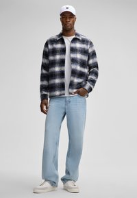 Kariertes Flanelljackett in Blau und Weiß, graues T-Shirt darunter, hellblaue weit geschnittene Jeans und weiße Sneakers. Eine weiße Mütze rundet den Look ab.