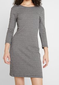 Dlouhé šaty s dlouhými rukávy v černo-bílém vzoru houndstooth. Elastická tkanina s lodní vardou a bočními rozparky na lemu.
