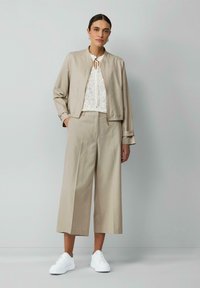 Beige bomberjack met ritsluiting, gecombineerd met beige broek met wijde pijpen en witte sneakers. Eronder een witte blouse met patroon.