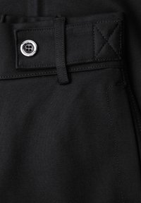 Nærbillede af sorte bukser med linning, sort bæltestrop, syet søm og sølvfarben firhullet knap på stofstrop.