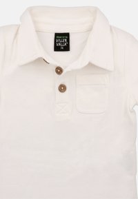Hvid økologisk bomulds polo-shirt med klassisk krave, tre brune knapper og en brystlomme med broderet detalje.