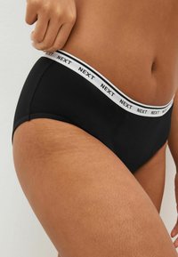 Zwarte slip met een witte elastische tailleband waarop de tekst "NEXT." staat. De stof ziet er glad en rekbaar uit, ontworpen voor comfort.
