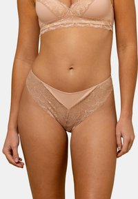 String en dentelle de couleur chair avec un bord festonné et un motif floral détaillé, fabriqué à partir d'un tissu doux et transparent avec un design minimaliste.