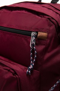 Sac à dos bordeaux en tissu texturé, doté d'une fermeture éclair noire, d'un accent en cuir et d'un cordon de tirage à motifs en bleu marine, rouge et blanc.