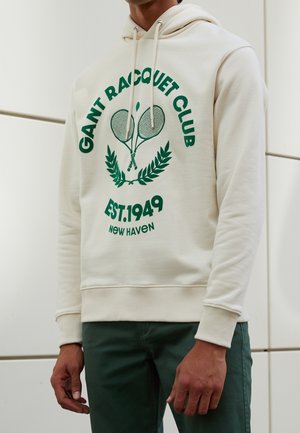 Beżowa bluza z kapturem z zielonym napisem i grafiką skrzyżowanych rakiet tenisowych, zestawiona z zielonymi spodniami, pokazana od szyi do połowy uda.