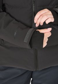 Veste isolante noire avec fermeture éclair et poignets élastiques avec ouverture pour le pouce, mettant en avant des matériaux texturés et un design épuré.