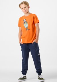 Orange bomulds T-shirt med hundegrafik, parret med navy blå cargo sweatpants med sidelommer. Hvide sneakers med grønne accenter.